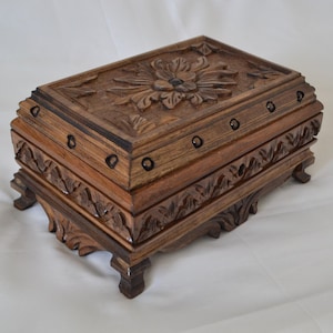 May include: A rectangular wooden box with intricate floral carvings on the lid and sides. The box is a warm brown color, with dark accents and small, dark circular details. It has four small feet.