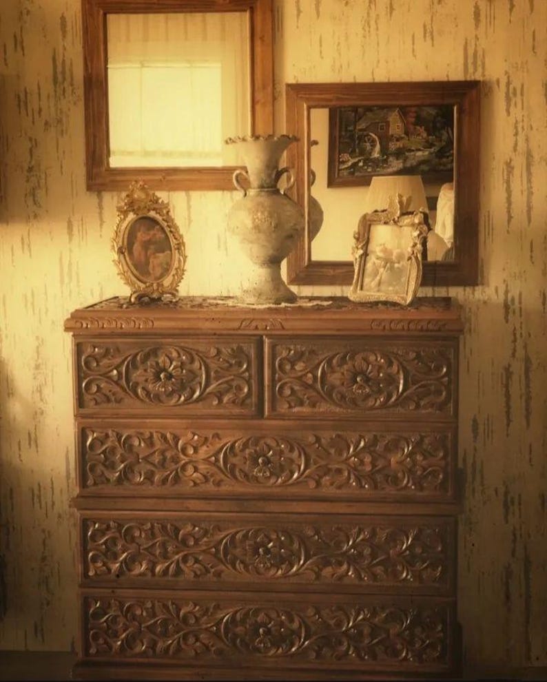 Puede incluir: Una c&oacute;moda de madera con cinco cajones, cada uno tallado intrincadamente con motivos florales. La c&oacute;moda est&aacute; decorada con un jarr&oacute;n, una imagen enmarcada y dos espejos.