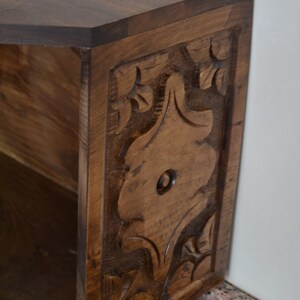 May include: A wooden side table with a dark brown finish. The side panel features a carved floral design. The table has a geometric top and a shelf inside. The table is made of solid wood.