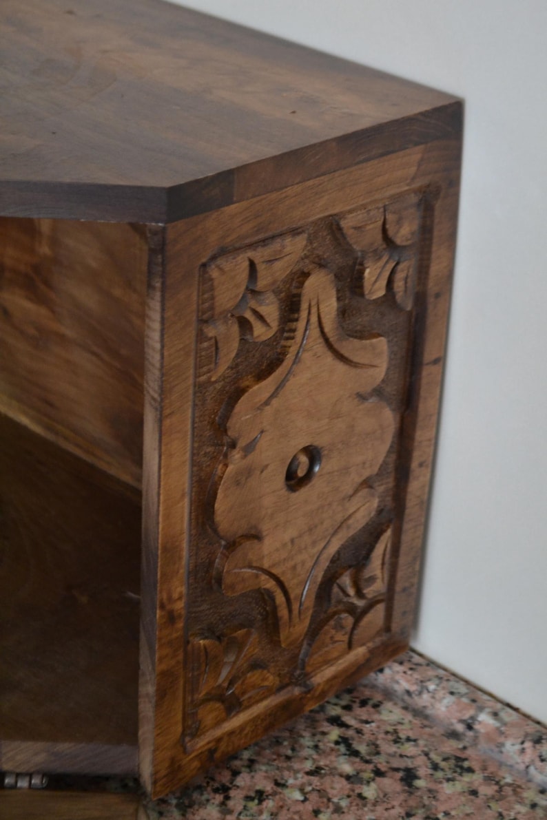 May include: A wooden side table with a dark brown finish. The side panel features a carved floral design. The table has a geometric top and a shelf inside. The table is made of solid wood.