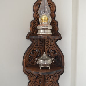 May include: A dark brown wooden corner shelf with intricate carvings. The shelf has two tiers and features a silver oil lamp with a glass chimney and a silver sugar bowl.