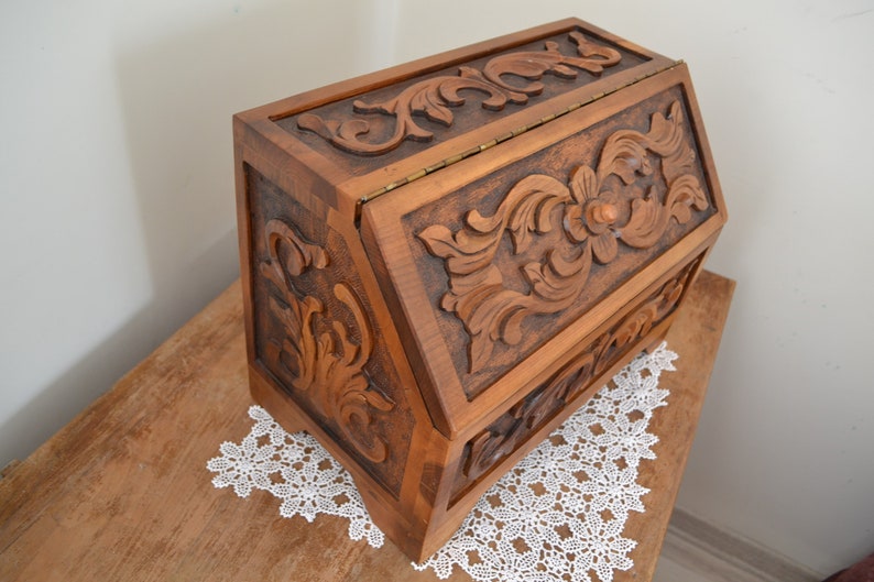 May include: A rectangular wooden box with a hinged lid, featuring intricate relief carvings of floral and scrolling leaf patterns on the front and side panels. The box is constructed from medium-brown wood and includes a small wooden knob on the lid.