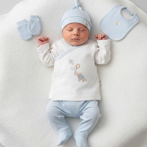 May include: A baby outfit set in light blue and white, including a long-sleeved wrap-style top with an embroidered elephant holding a kite, matching footed trousers, a knotted beanie hat, a pair of mittens, and a small bib with a matching logo tag.