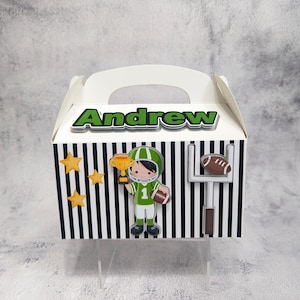 Puede incluir: Una caja de regalo blanca con un diseño de rayas blancas y negras. La caja tiene un asa y presenta un gráfico de un jugador de fútbol verde y blanco, un trofeo dorado, un balón de fútbol y una portería. El nombre "Andrew" está impreso en la parte delantera de la caja.