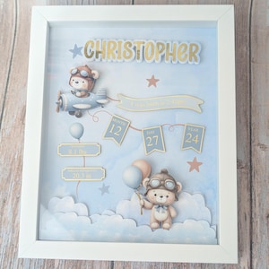 May include: A framed baby announcement with a blue and white background. The announcement includes the name "CHRISTOPHER" in gold lettering, a teddy bear in a pilot's hat, and the baby's birth details: 8.1 lbs weight, 20.3 inches in length, and the date of birth.