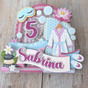 May include: A layered paper craft cake topper with a spa theme. Features a pink and white colour scheme, a number 5, a bathrobe, slippers, and the name "Sabrina". Includes a bathtub, bubbles, and a lotus flower.