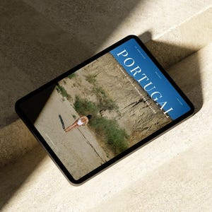 May include: A tablet computer displaying a magazine cover with the word "PORTUGAL" in large blue letters. The cover features a photo of a person in a white swimsuit standing on a sandy beach.