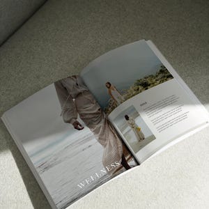 May include: Open magazine with the word "WELLNESS" on the cover. The magazine features a woman in a flowing white dress walking on a beach and another woman in a white dress standing on a cliff.