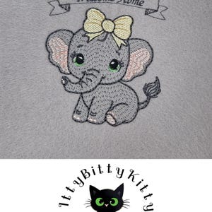 Puede incluir: Diseño bordado de un elefante gris con un lazo amarillo, sentado sobre un fondo gris. La pancarta sobre el elefante dice "Welcome Home".