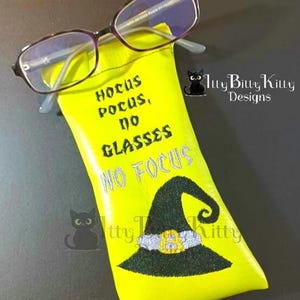May include: A bright yellow glasses case with the text "Hocus Pocus, No Glasses, No Focus" and a witch hat design. The case holds a pair of eyeglasses with purple frames. A black cat logo is visible.