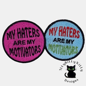 May include: Two round embroidered patches. One is pink with black text that reads "MY HATERS ARE MY MOTIVATORS." The other is light blue with red and green text that reads the same. A black cat logo is in the bottom right.