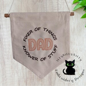 May include: A beige fabric banner with the words "FIXER OF THINGS" and "KNOW OF STUFF" embroidered in black around the word "DAD" in orange. A black cat with green eyes is in the lower right corner. The banner is suspended from a wooden dowel.