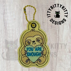 May include: A yellow embroidered keychain featuring a brown sloth holding a light green heart. The heart has the text YOU ARE ENOUGH stitched in blue thread. The keychain includes a yellow ball chain for attachment and has a die-cut shape.