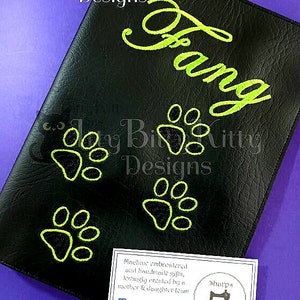 Könnte beinhalten: Ein schwarzes Ledernotizbuch mit dem Namen "Fang", der in grünem Garn gestickt ist. Das Notizbuch ist mit grünen Pfotenabdrücken verziert. Eine Visitenkarte für "Sharp Stitches" befindet sich in der unteren rechten Ecke des Bildes.