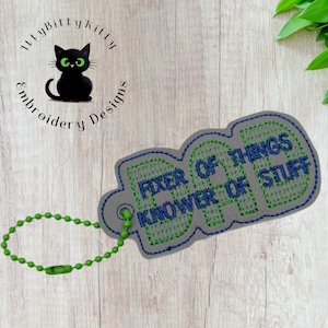 May include: A grey embroidered keyring with the text "FIXER OF THINGS, KNOWER OF STUFF" in green and blue stitching. The keyring has a green beaded chain and a small black cat logo.
