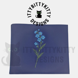 May include: A rectangular dark blue leather-like folder featuring an embroidered design of a single stem with light blue flowers and green leaves. Above the folder, the text ITTYBITTYKITTY DESIGNS is printed in a circular black font.