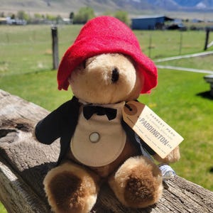 May include: A Paddington Bear plush toy wearing a red hat, black jacket, and bow tie. The bear has a tag that reads "Darkest Peru to London, England". The plush is tan and brown.