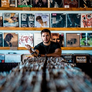 May include: A man wearing a black shirt and a silver necklace sits in front of a wall of vinyl records. The records are displayed on shelves with wooden frames. The man is holding a record in his hand.