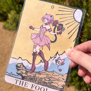 Op de afbeelding: Een tarotkaartillustratie met een personage in een roze jurk en laarzen, met kattenoren en een staart. De kaart is gelabeld "THE FOOL" en toont een zon, bergen en een klein wezen. De kaart wordt in een doorzichtige plastic hoes gehouden.