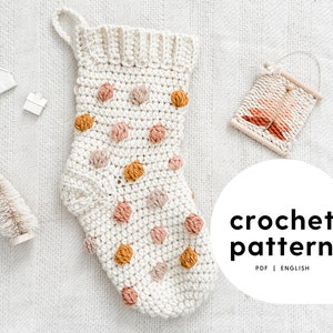 May include: A crochet pattern for a white stocking with pink, brown, and yellow polka dots. The pattern is available as a PDF in English. The image also includes a small white Christmas tree, a white house, and a woven wall hanging.