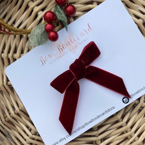 Può includere: Un fiocco per capelli in velluto bordeaux con un semplice design a nodo. Il fiocco è su una carta bianca con il testo "Bow Boulevard Handmade Hair Bows" e un indirizzo di sito web.