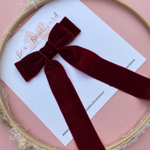 Puede incluir: Un lazo de terciopelo burdeos con colas largas, mostrado en una tarjeta blanca con el texto "Bow Boulevard Hand Made Hair", y un aro de bordado de madera con flores blancas secas.