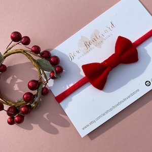 Può includere: Fascia per capelli con fiocco in velluto rosso e fascia elastica rossa. La fascia è su una carta bianca con il testo "Bow Boulevard Handmade Hair Bows" e l'indirizzo del sito web "www.etsy.com/ie/shop/bowboulevardstore".