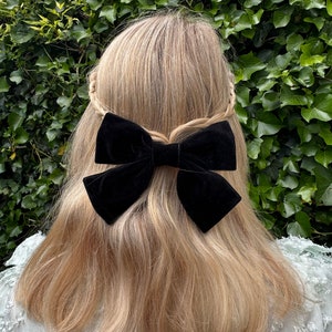 Pode incluir: Um acessório de cabelo com laço de veludo preto é preso na parte de trás do cabelo loiro. O cabelo é penteado com uma trança na coroa. O fundo é uma sebe verde exuberante.