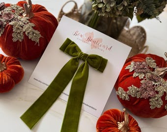 May include: An olive green velvet hair bow with long streamers rests on a white card with the text "Bow Boulevard Handmade Hair Bows". The bow is surrounded by small, orange velvet pumpkins with gold stems and decorative accents.