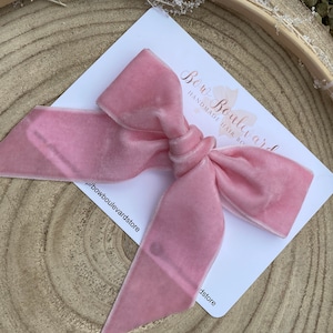 May include: A pale pink velvet hair bow with long tails, knotted in the centre, displayed on a white card with the text "Bow Boulevard Handmade Hair Bows". The bow is on a wooden surface.
