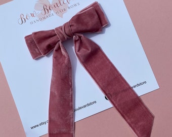 May include: A dusty rose velvet hair bow with long, flowing tails. The bow is centered on a white card with the text "Bow Boulevard Handmade Hair Bows" and the Etsy store URL. The background is a solid, soft pink.