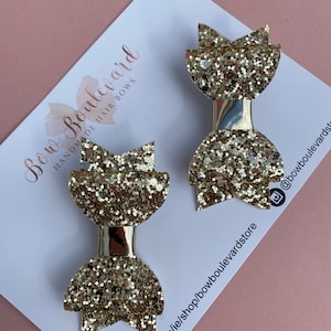 May include: Two gold glitter hair bows with a gold metallic centre. The bows are on a white card with the text "Bow Boulevard Handmade Hair Bows" and a website address.