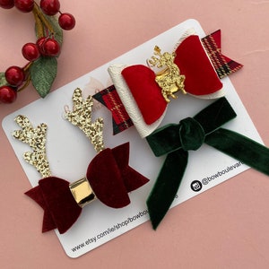 Könnte beinhalten: Drei Weihnachts-Haarspangen. Eine ist aus rotem Samt mit goldenen Glitzer-Geweih, eine ist weiß mit einer roten Samt Schleife und einem goldenen Rentier, und eine ist aus grünem Samt.