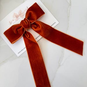Puede incluir: Un lazo de terciopelo naranja quemado con largas colas fluidas. El lazo está atado en el centro y descansa sobre una tarjeta blanca con el texto "Bow Boulevard" y "www.etsy.com". El accesorio es un lazo para el pelo.