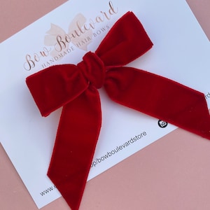 May include: A large, red velvet hair bow. The bow is tied in a classic, symmetrical shape with pointed ends. The bow is on a white background with the text "Bow Boulevard Handmade Hair Bows" and a website address.