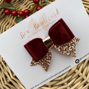 Puede incluir: Un lazo para el pelo de terciopelo burdeos con detalles de purpurina dorada y un centro dorado. El lazo está sobre una tarjeta blanca con el texto "Bow Boulevard Handmade Hair Bows". La tarjeta también incluye una dirección web y un identificador de Instagram.