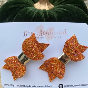 Puede incluir: Dos lazos para el pelo de color naranja con purpurina y ribete dorado. Los lazos están sobre una tarjeta blanca con el texto "Bow Boulevard Made Hair Bows" y la dirección del sitio web "www.etsy.com/ie/shop/bowboulevardstore" y el nombre de usuario de Instagram "@bowboulevardstore".