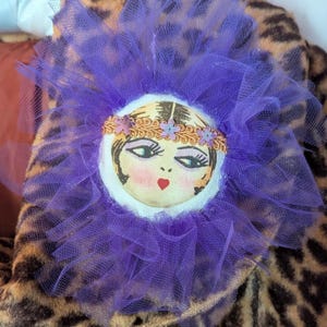 May include: A circular fabric brooch featuring a printed illustration of a face with dark hair, defined eyes, and a red heart-shaped mouth. The center is surrounded by a ruffled purple tulle trim and accented with a gold braided headband with small flowers.