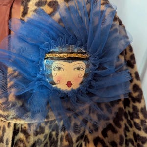 May include: A circular brooch featuring a hand-painted face with dark hair, red lips, and a gold headband, surrounded by a ruffled blue tulle trim. The brooch is pinned to a garment with a brown and black leopard print pattern.