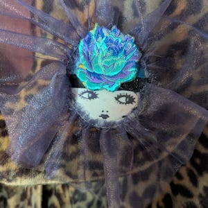 May include: A leopard print fabric accessory featuring a ruffled purple sheer tulle collar. Centered on the collar is a vibrant embroidered flower in shades of teal, blue, and purple, positioned above a small white patch with a printed face illustration.