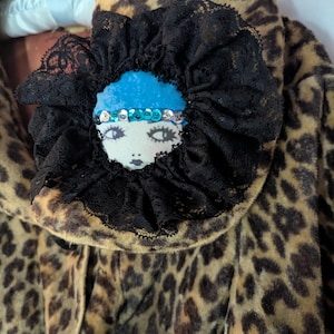 May include: A leopard print garment featuring a circular brooch with a black lace ruffle. The brooch displays a stylized face with a blue headpiece, a sequined band, and dark eyes. The garment has a soft, textured surface and a white ribbon detail.