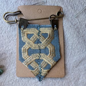 May include: A shield-shaped fabric brooch featuring a blue linen base with intricate gold-colored thread embroidery in a geometric knot pattern. The fabric is attached to a bronze-toned metal safety pin by two small metal clasps at the top corners.