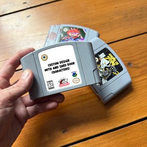 May include: Three gray Nintendo 64 game cartridges. One cartridge has a white label with the text "CUSTOM DESIGN WITH AND SHED DOOR CHARACTERS!". The other cartridges have colorful graphics. The cartridges are on a wooden surface.