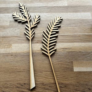 Op de afbeelding: Twee houten tarwestengels met gedetailleerde bladerpatronen. De stengels zijn lichtbruin en lijken lasergesneden te zijn. Ze liggen op een houten oppervlak.
