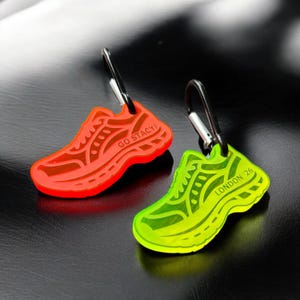 Puede incluir: Dos llaveros con forma de zapatilla. Uno es de color naranja neón con el texto "GO STACY!" y el otro es verde neón con el texto "LONDON '26". Ambos llaveros tienen un mosquetón plateado.