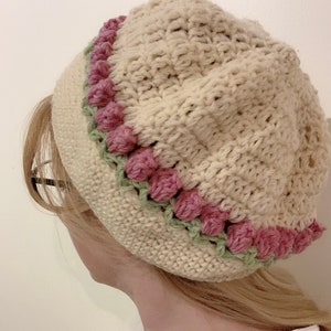 May include: A crocheted beanie hat in cream with a pink and green crocheted trim. The trim features a row of pink pom-poms.