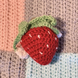 Pode incluir: Um morango de croch&ecirc; feito &agrave; m&atilde;o, com folhas verdes e sementes brancas. O morango &eacute; vermelho e est&aacute; sobre um fundo de patchwork colorido de quadrados rosa, cinza e azul. O trabalho de croch&ecirc; &eacute; detalhado.