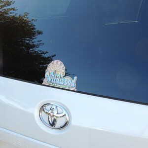 May include: A white car with a sticker on the rear window. The sticker is a colourful design of a Ferris wheel and the words "Wildwood, Life in a Sandcastle".
