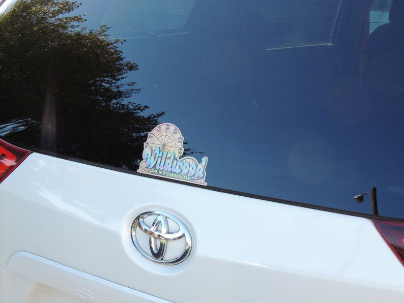 May include: A white car with a sticker on the rear window. The sticker is a colourful design of a Ferris wheel and the words "Wildwood, Life in a Sandcastle".