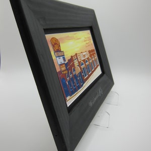 May include: A black wooden frame with a vintage postcard inside. The postcard shows a building with the text "Uncle Lou's Restaurant" on the front. The frame is on a white surface with two clear acrylic stands.